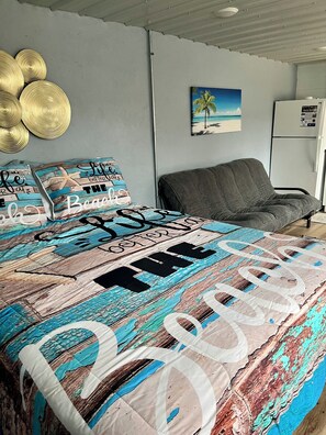 Un dormitorio con una cama, un sillón gris, un cuadro en la pared y un refrigerador.