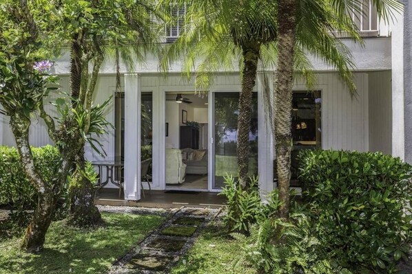 A modern house with a covered patio, a visible living room with a sofa, and a garden with various plants and a palm tree. - Princeville: Enjoy Amazing Sunsets & Views of Ocean 1 BD Condo With Pullout Sofa (Princeville)