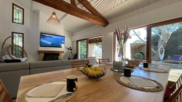 A spacious living room with a wooden dining table, a television, and a large window offering a view of the outdoors. - la villa familiale la palmyre (Les Mathes)