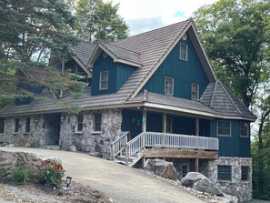 A two-story stone house with a steeply pitched roof, a front porch with a railing, and a stone foundation.