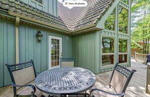 Une terrasse couverte avec une table ronde en métal et quatre chaises, une porte et des fenêtres donnant sur une zone boisée.