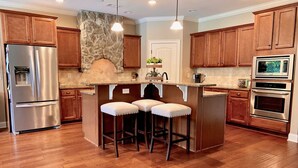 A modern kitchen with wooden cabinets, a stone backsplash, stainless steel appliances, and a central island with bar stools. - Beautiful 5 BR, 3 BA, home only minutes away from the Masters and Peach Jam! (North Augusta)