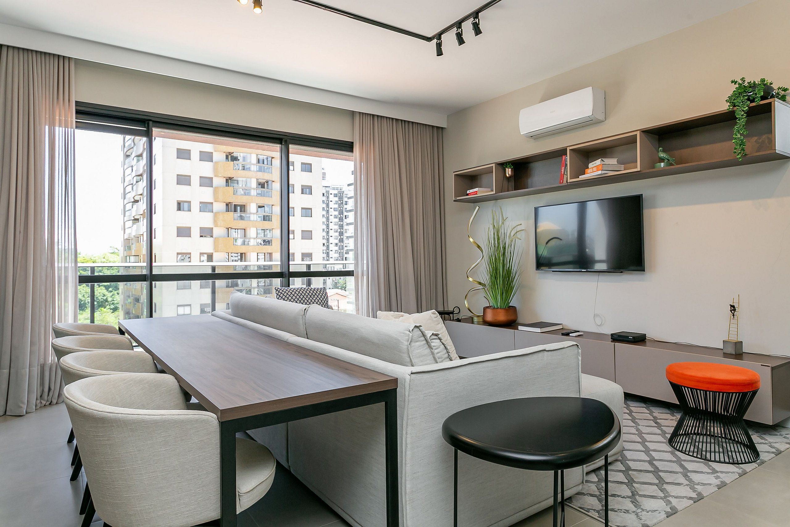 Een moderne woonkamer met een bank, eettafel en stoelen, een aan de muur bevestigde televisie en een groot raam met gordijnen.