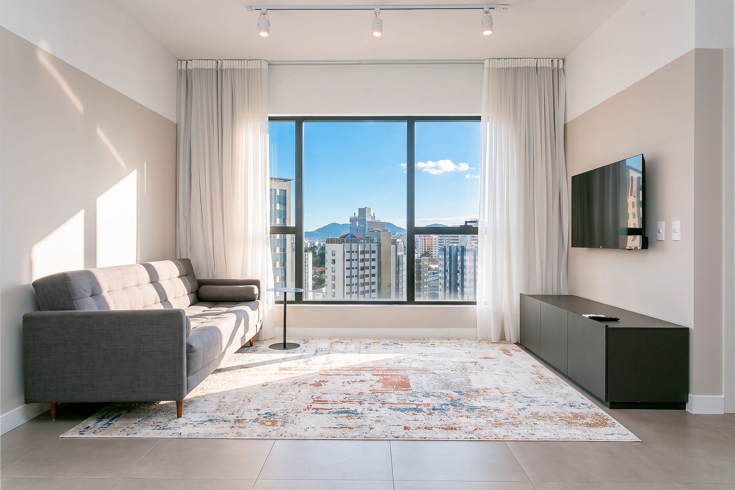 A modern living room with a sofa, a coffee table, a TV mounted on the wall, and a large window offering a city view.