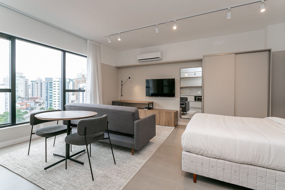 A modern hotel room with a large window, a sofa, a bed, a TV, and a small table.