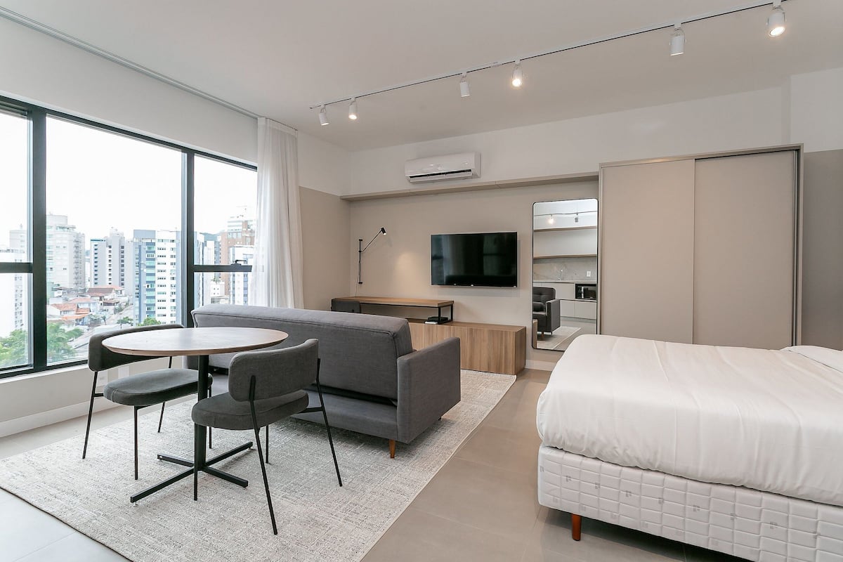 Quarto de hotel moderno com uma janela ampla, um sofá, uma cama, uma televisão e uma pequena mesa.