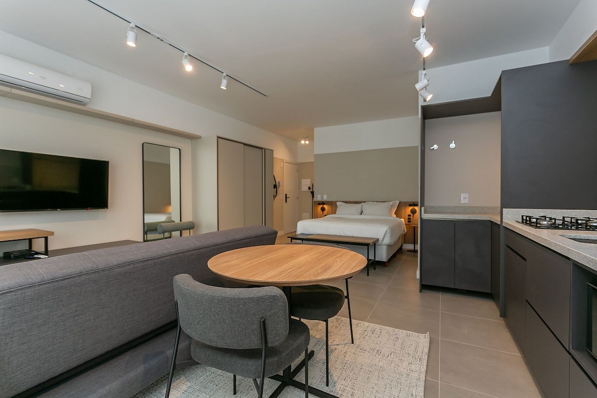 Um espaço de estar moderno e compacto com sofá, uma mesa redonda de madeira, TV na parede, cama com mesa de cabeceira e cozinha.