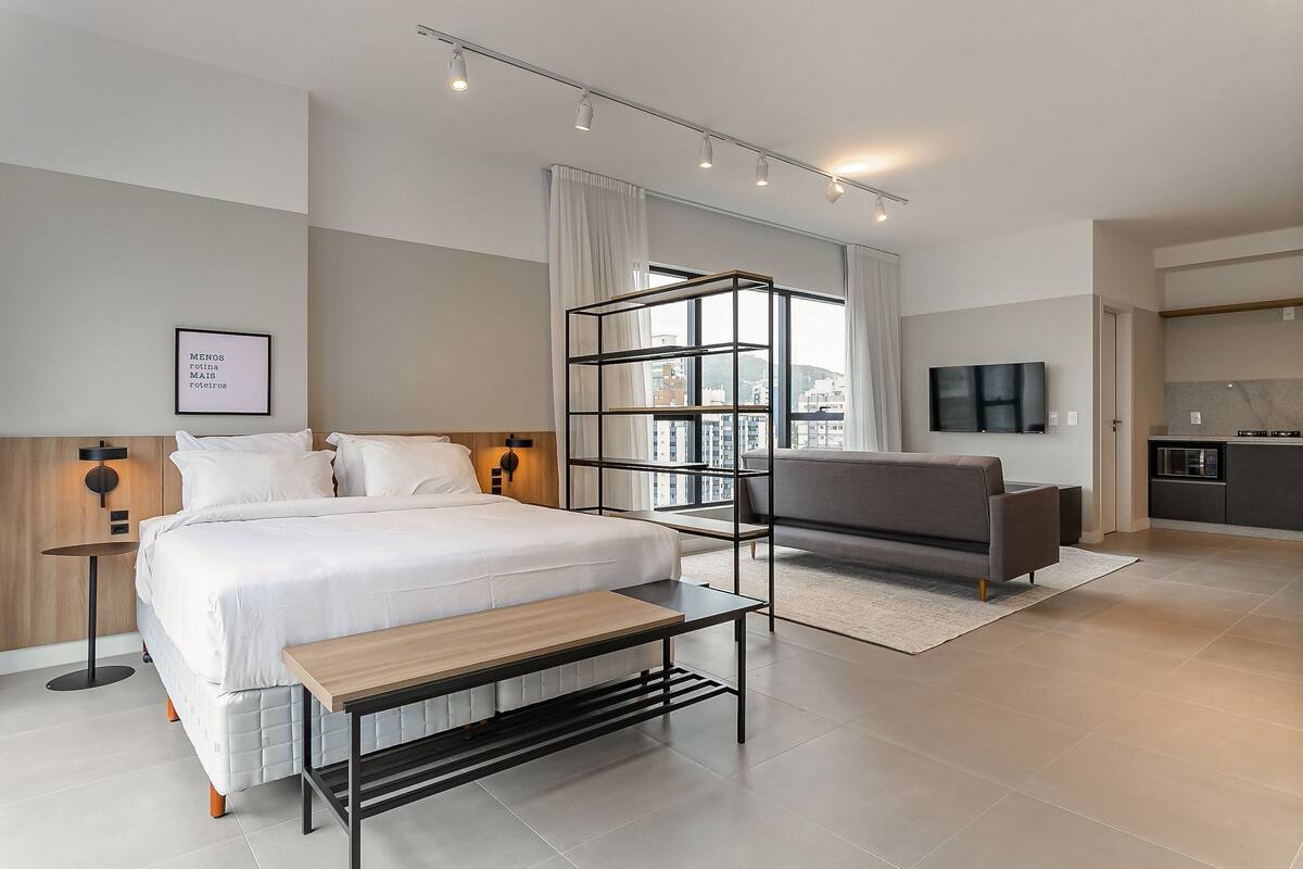 Quarto de hotel moderno com uma cama grande, um sofá, uma mesinha, uma televisão e uma cozinha.