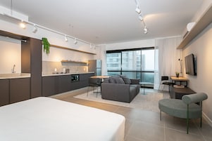 A modern hotel room with a bed, kitchenette, seating area, and a large window.