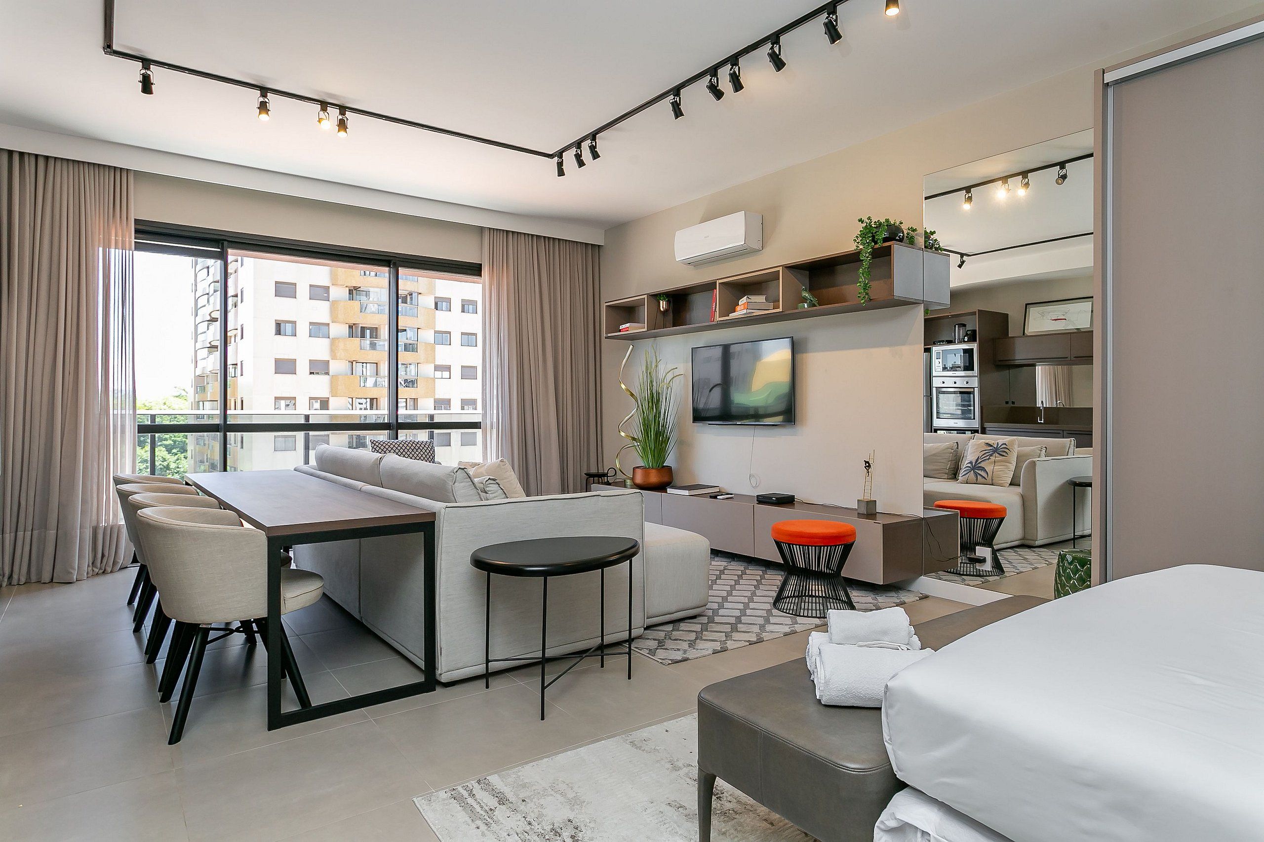 A modern hotel room with a sofa, dining table, and chairs, a small kitchen area, a TV, and a large window with curtains.