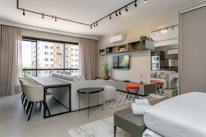 Et moderne hotelværelse med sofa, spisebord, stole, et lille tekøkken, fjernsyn og et stort vindue med gardiner.