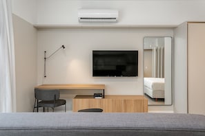 A modern hotel room with a flat-screen TV mounted on the wall, a desk with a chair, a bedside table, and a large mirror reflecting the bed. - Studios Tower by Viva - Prédio com piscina no rooftop no centro de Floripa (Florianópolis)