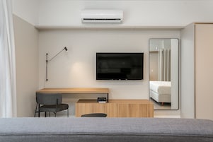 Una moderna camera d'albergo con una TV a schermo piatto fissata al muro, una scrivania con una sedia, un comodino e un grande specchio che riflette il letto.