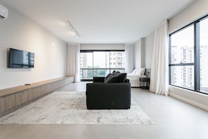 Un soggiorno moderno con un divano nero, una televisione a schermo piatto, un'ampia finestra con tende e vista su edifici alti.