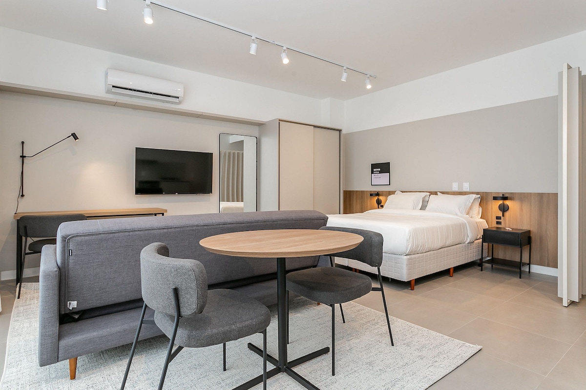 A modern hotel room with a sofa, a round table, a bed, a TV, and a bedside table with a lamp.