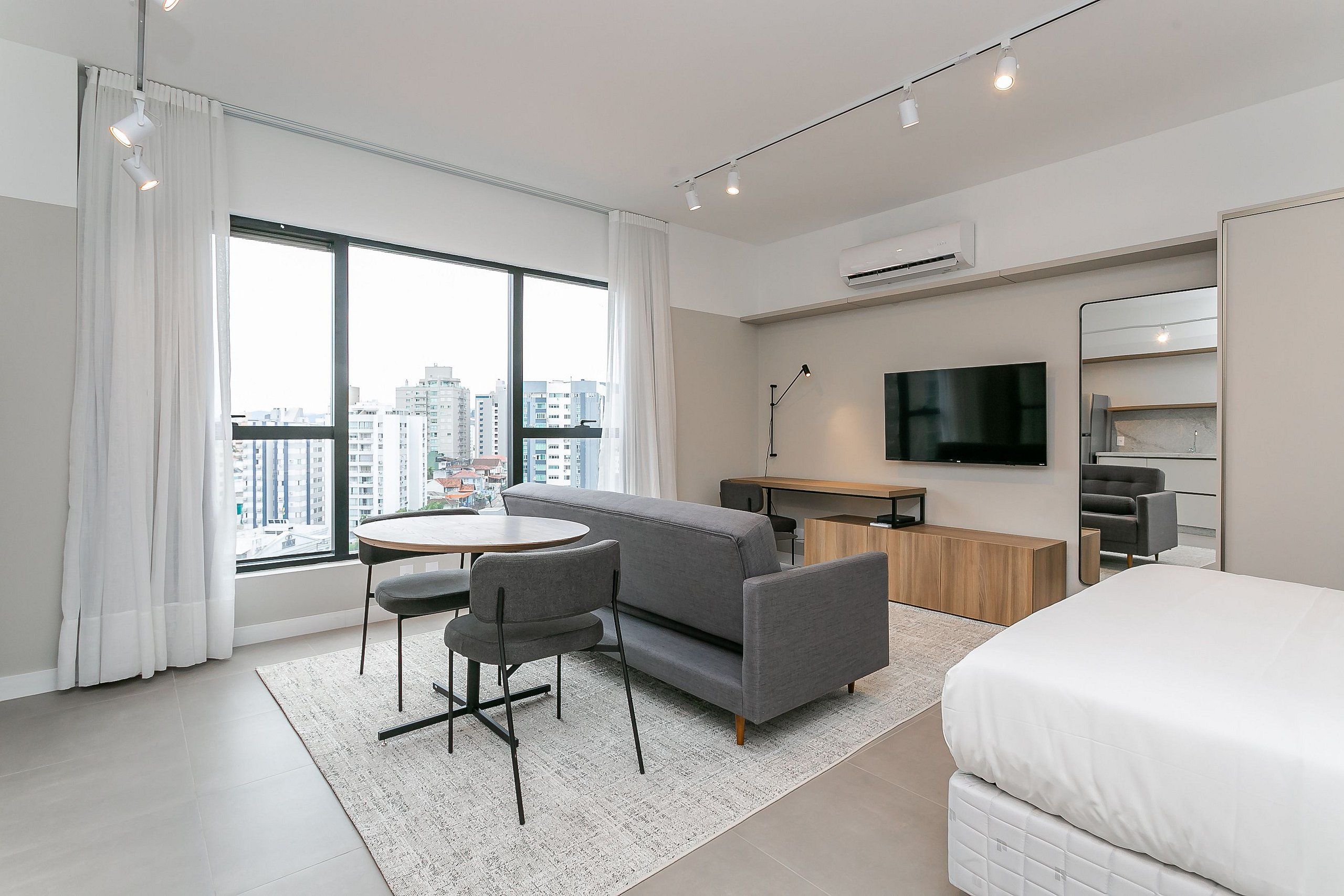 A modern hotel room with a large window, a sofa, a dining table with chairs, a TV, and a bed.