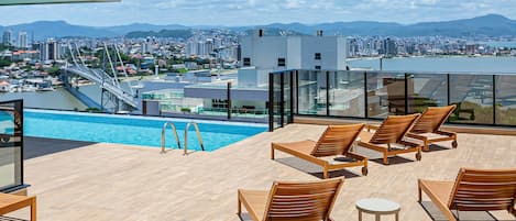 A rooftop pool area with wooden lounge chairs and a view of a cityscape and mountains.