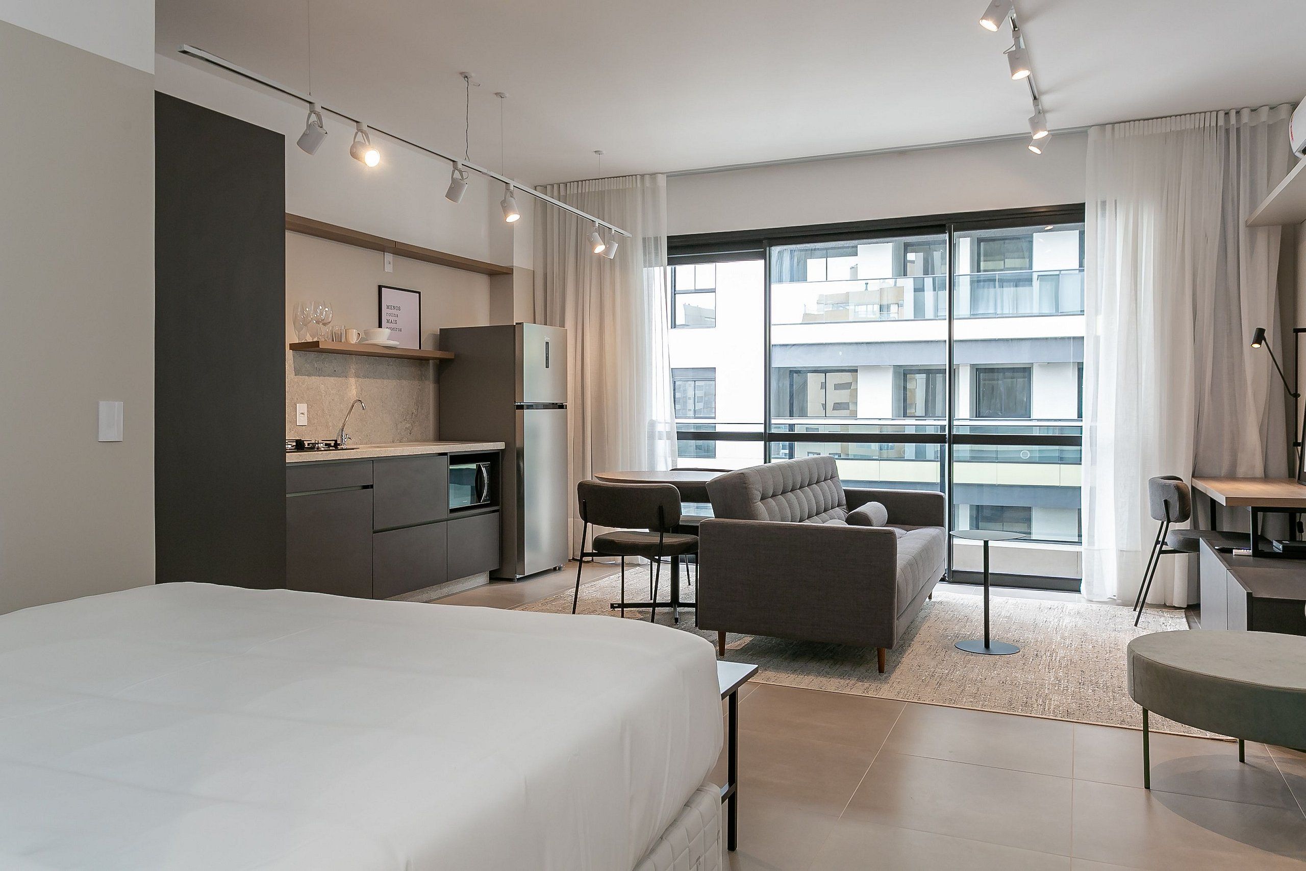 A modern hotel room with a large bed, a dining area with a table and chairs, a sofa, a kitchen area, and a large window with a city view.