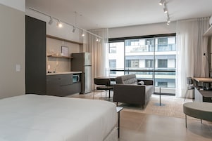 A modern hotel room with a large bed, a dining area with a table and chairs, a sofa, a kitchen area, and a large window with a city view.