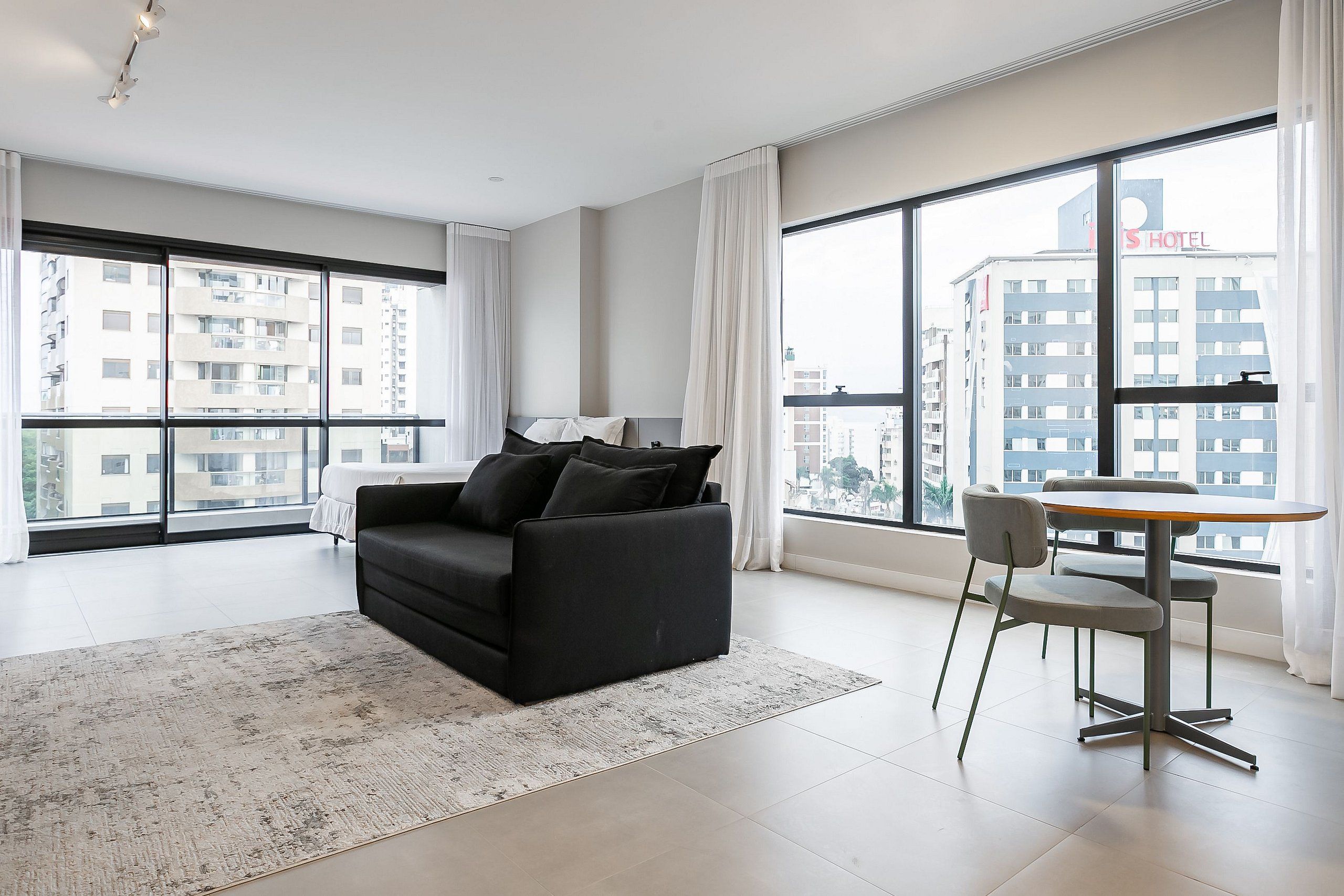 Une chambre d’hôtel moderne avec une grande fenêtre, un canapé noir, une table à manger ronde et une chaise grise.