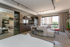 A modern hotel room with a large bed, a dining area, kitchenette, and a balcony with a view of the city.