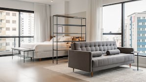 A modern bedroom with a grey sofa, a bed with white bedding, a black metal shelf unit, and large windows offering a city view. - Studios Tower by Viva - Prédio com piscina no rooftop no centro de Floripa (Florianópolis)