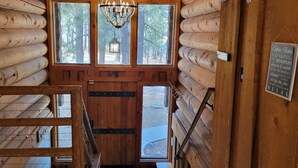 L’intérieur d’une cabane en rondins, avec un escalier en bois, de grandes fenêtres donnant sur la forêt et un lustre.