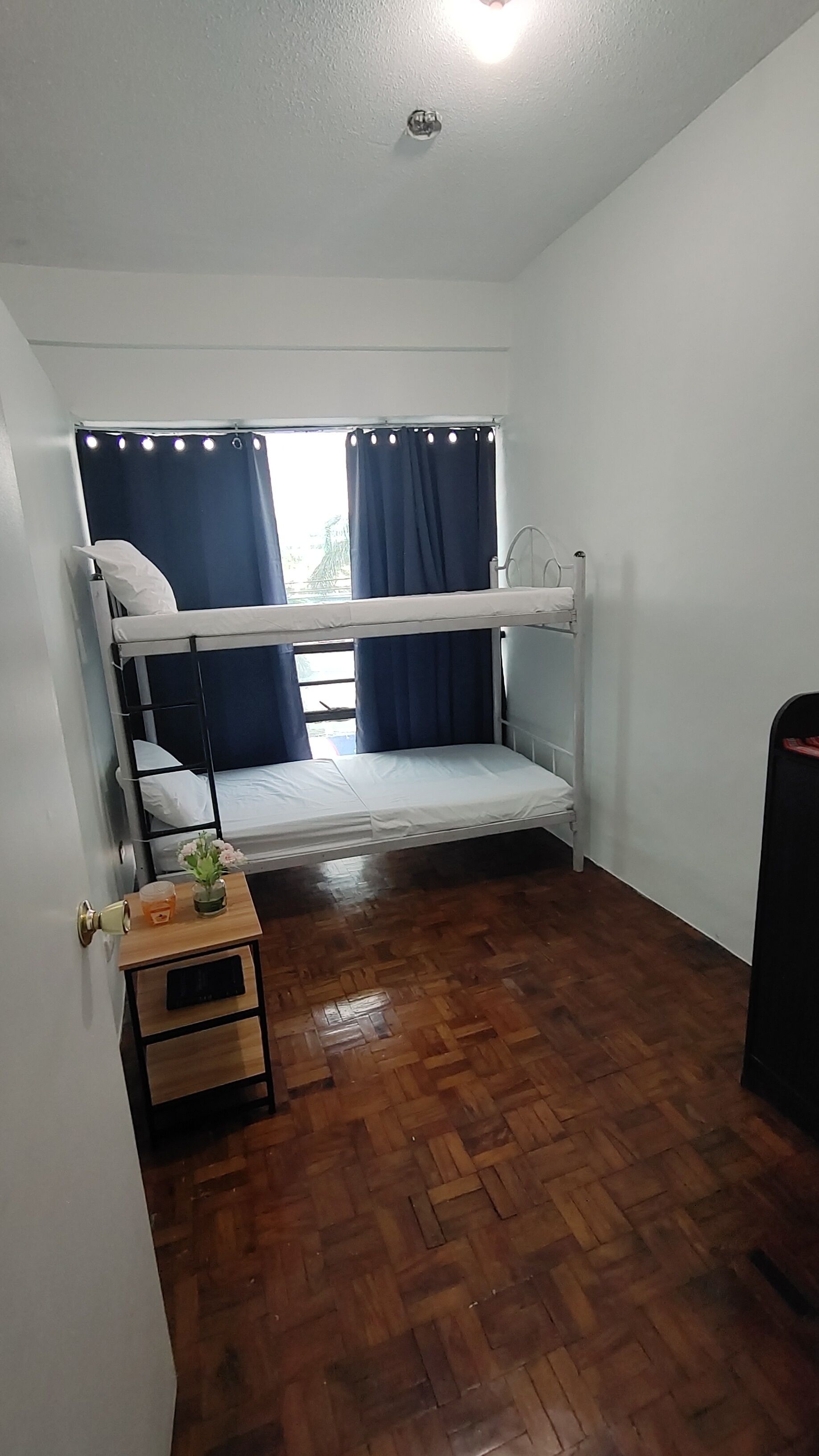 Una habitación pequeña con una sola cama, piso de madera, ventana con cortinas, mesita de noche con una planta y un radiador.