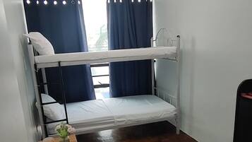Una habitación pequeña con una sola cama, piso de madera, ventana con cortinas, mesita de noche con una planta y un radiador.