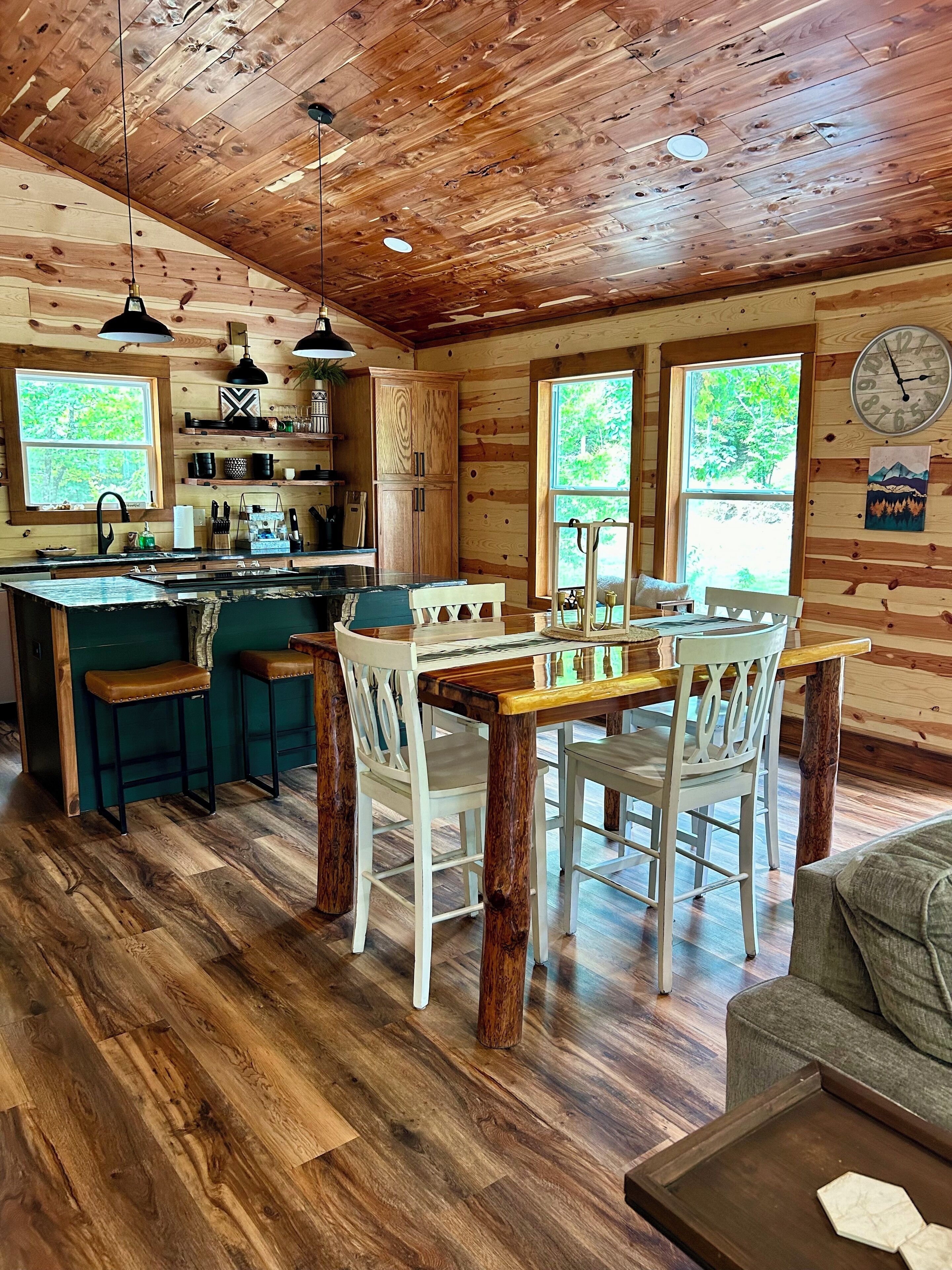 A rustic cabin interior with a wooden ceiling, a kitchen with a central island, and a dining area with a table and chairs.