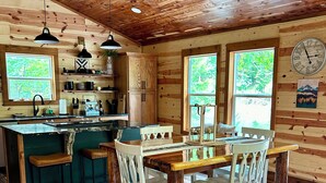 A rustic cabin interior with a wooden ceiling, a kitchen with a central island, and a dining area with a table and chairs.