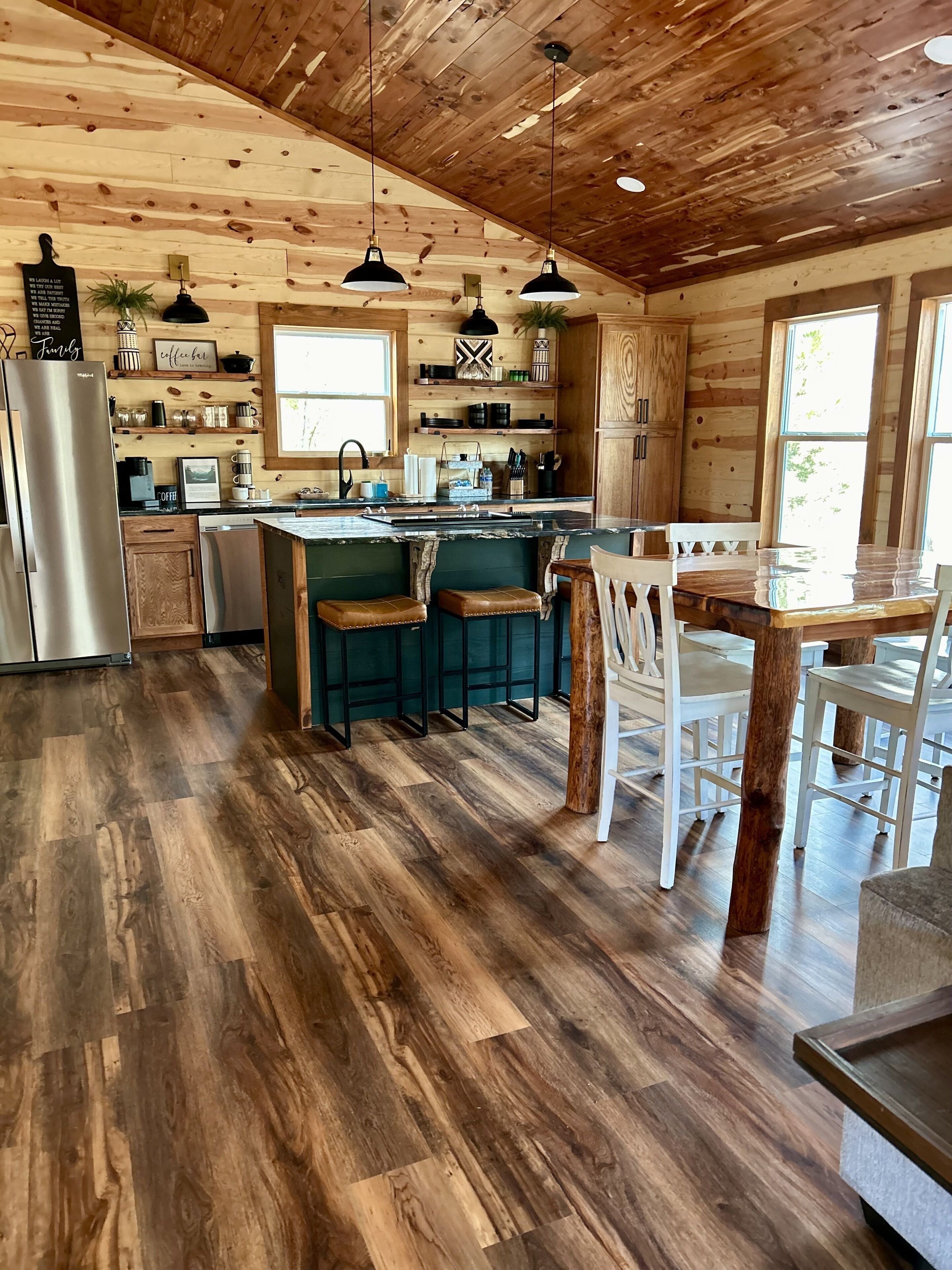 A rustic kitchen with wooden walls, a central island with stools, and a stainless steel refrigerator.