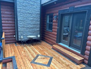 Una cabaña de troncos con pared de piedra, una terraza de madera, una mesa con sillas y un gran ventanal corredizo.