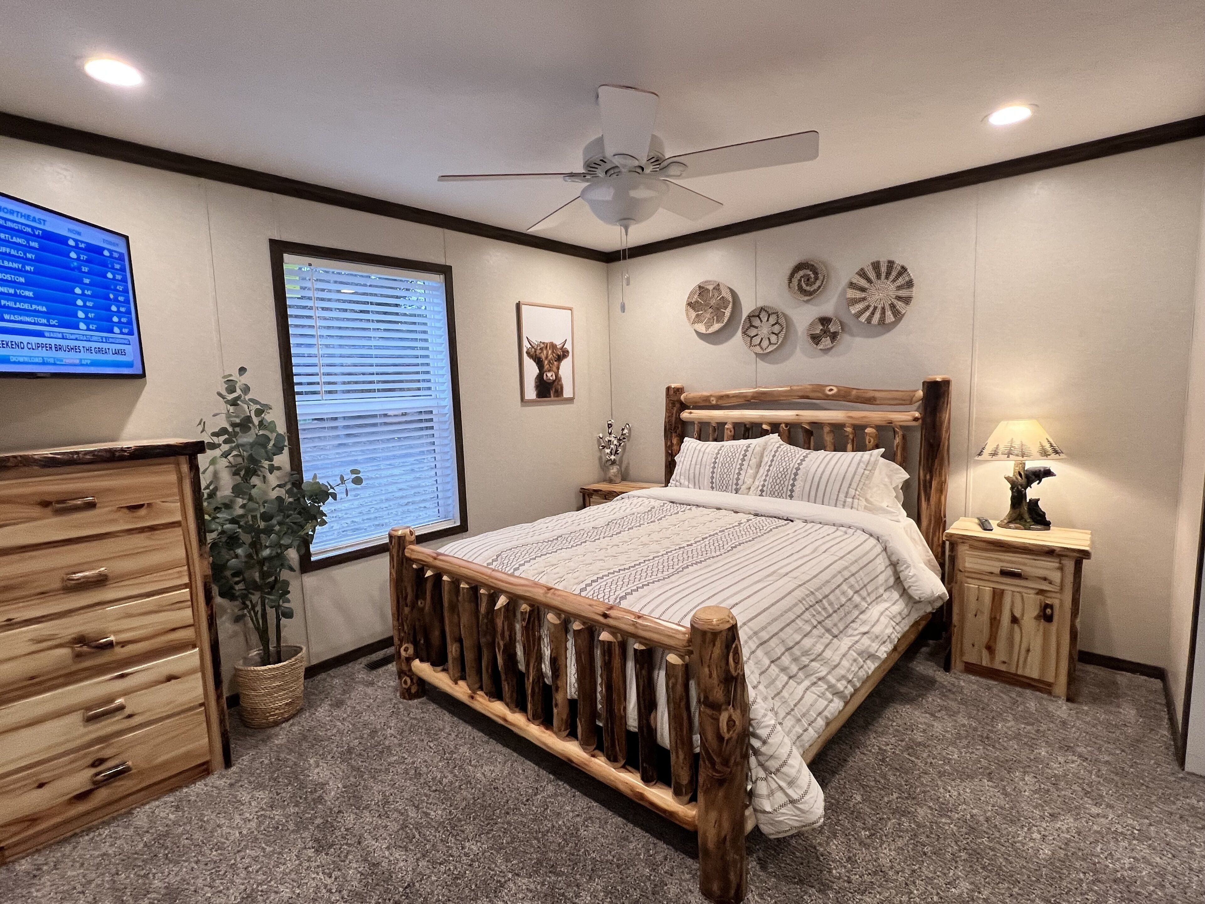 Une chambre à coucher avec un lit en bois, une table de chevet, une commode, un ventilateur de plafond et un téléviseur fixé au mur qui affiche les prévisions météorologiques.