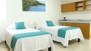 A room with two beds, a kitchen area, and a painting of a coastal scene.