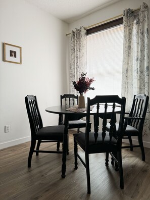 A dining area with a round table, four chairs, a vase with flowers, and a window with curtains.