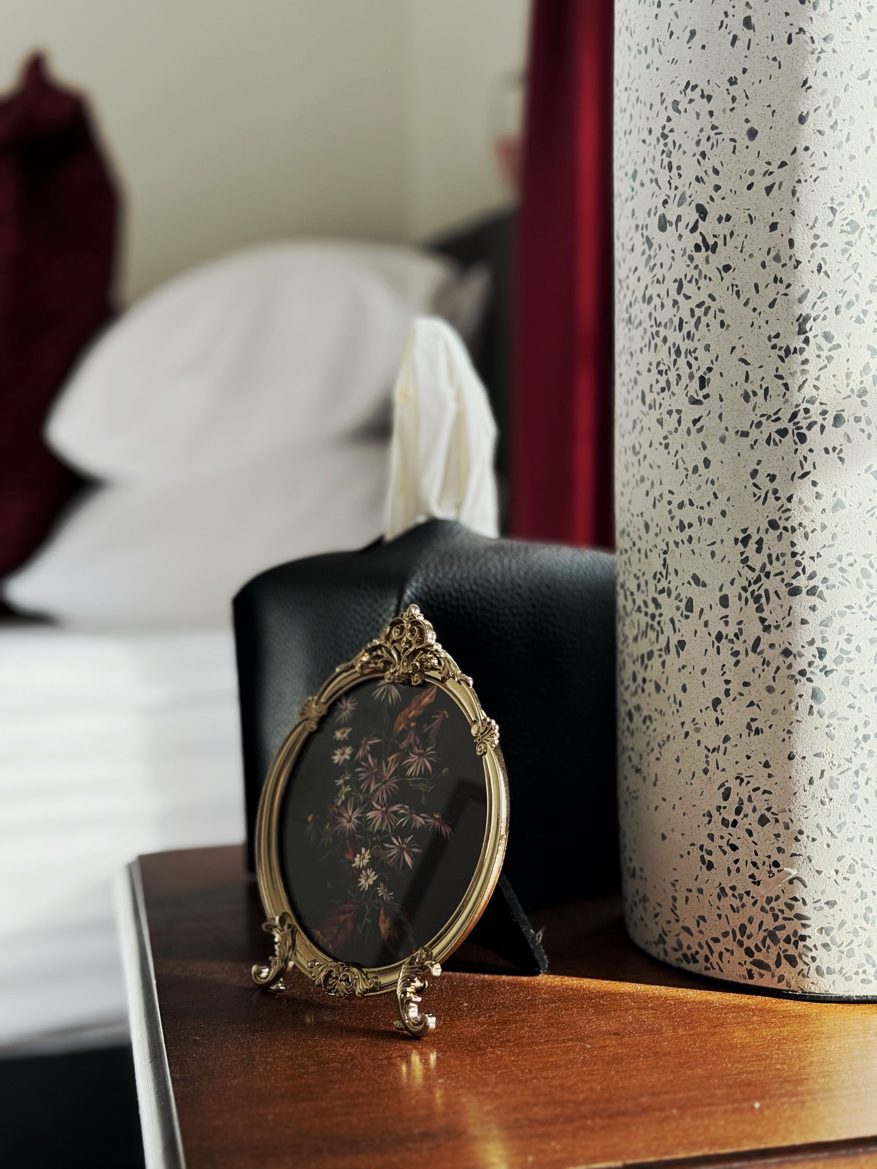A framed picture on a wooden surface, a white textured vase, and a black object in the background.
