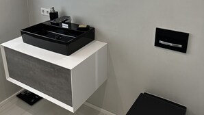 A modern bathroom with a white vanity, a black sink, a mirror with lights, and a black trash bin. - Vollausgestattete Wohnung im Neubau (Waldshut-Tiengen)