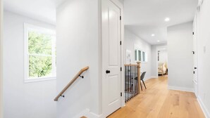 Un pasillo con escalera de madera, paredes blancas y vista a una habitación con cama y ventana.