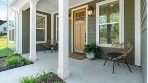 Una casa con porche, dos sillas de mimbre, una puerta de madera y una planta en maceta.