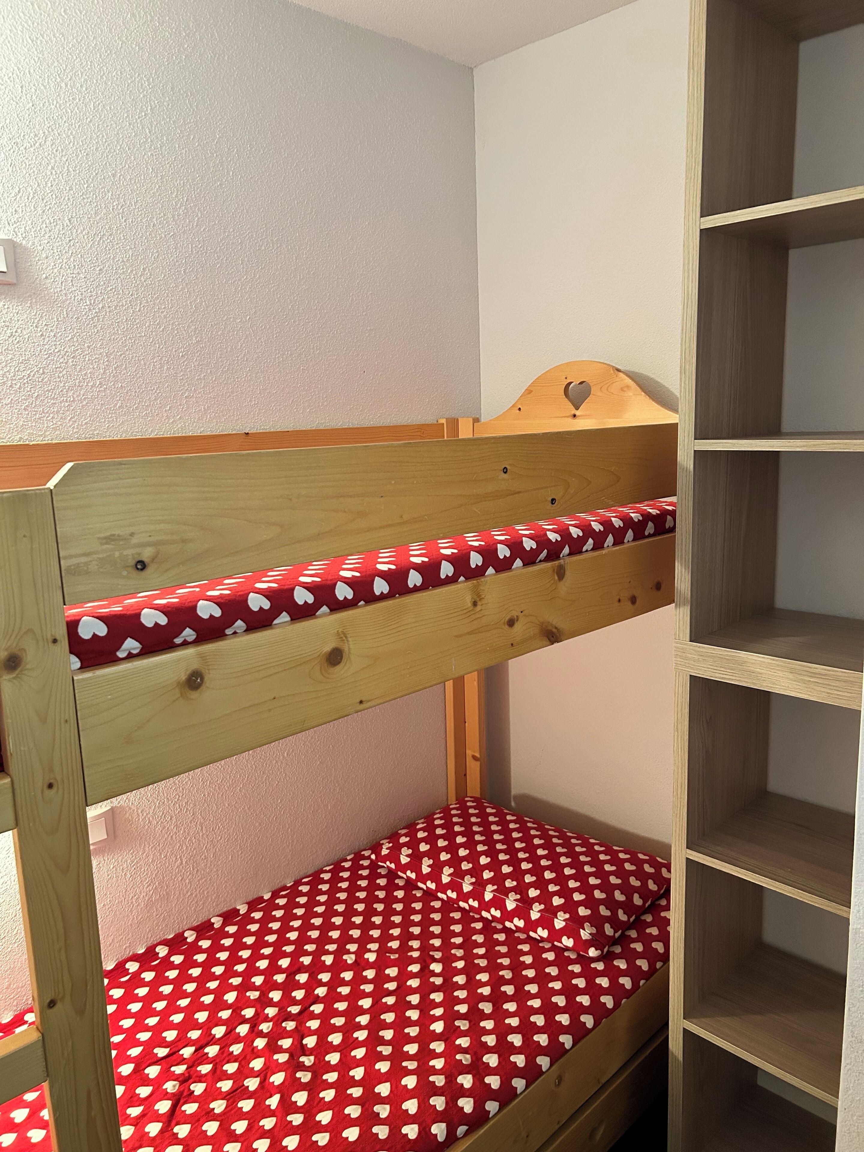 A bunk bed with a red mattress featuring a heart pattern, wooden headboard, and a wooden shelf unit beside it.
