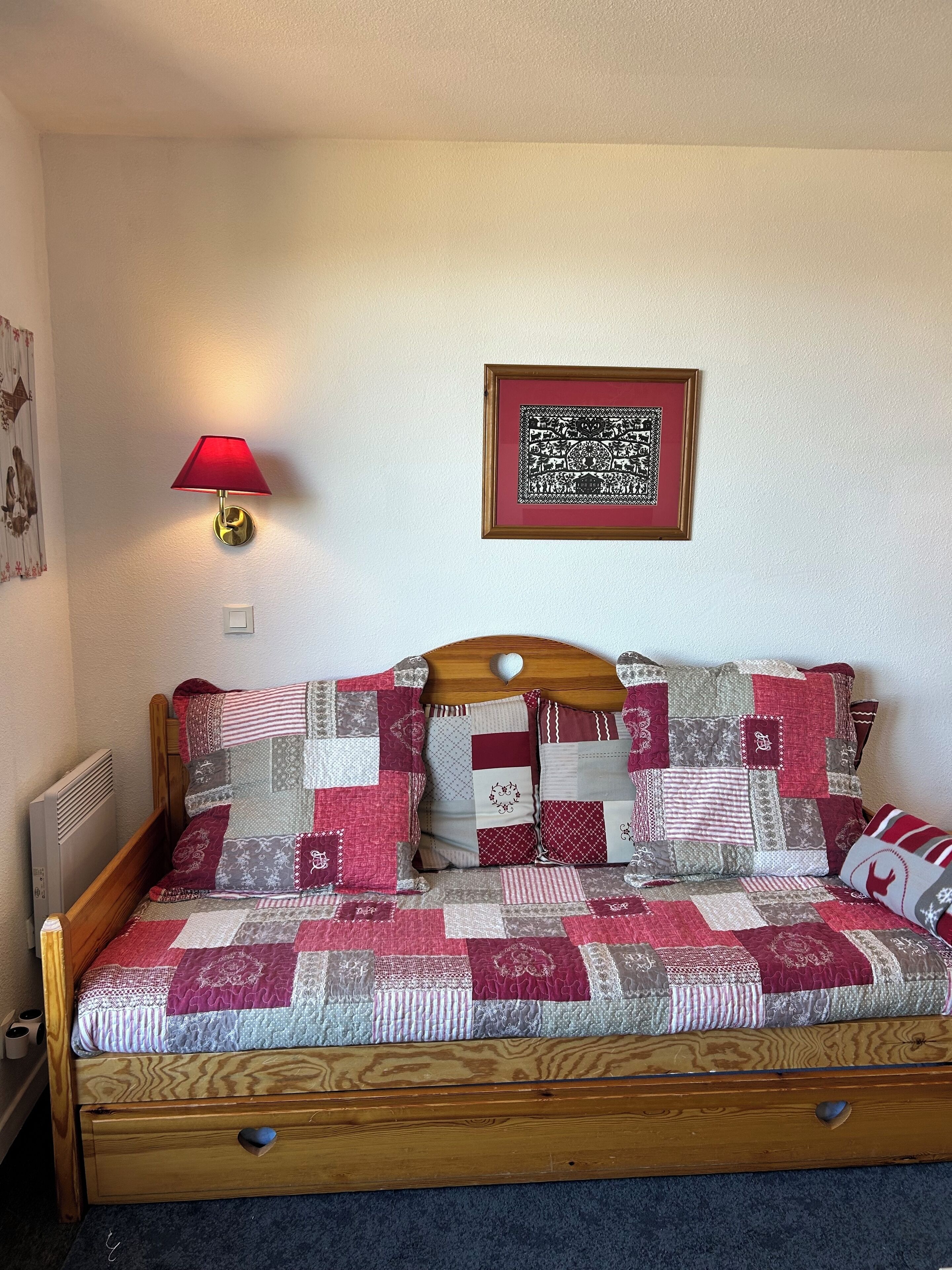 Un lit avec un cadre en bois, une lampe rouge fixée au mur et un tableau encadré au-dessus.