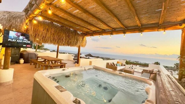 Un espace avec un jacuzzi, un écran de télévision, des meubles en bois et une vue sur l’océan au coucher du soleil.