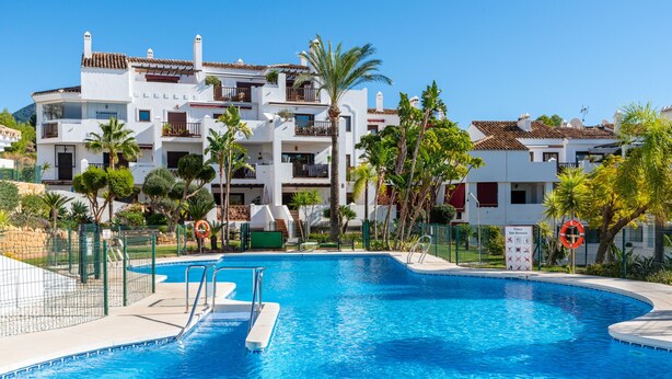 Una piscina con agua cristalina y azul, rodeada por una valla y contigua a un edificio de varios pisos con balcones y palmeras.