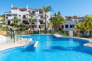 A swimming pool with a clear blue water, surrounded by a fence, and adjacent to a multi-story building with balconies and palm trees. - Finca San Antonio | Apartment with pool views near Mijas (Mijas Pueblo)