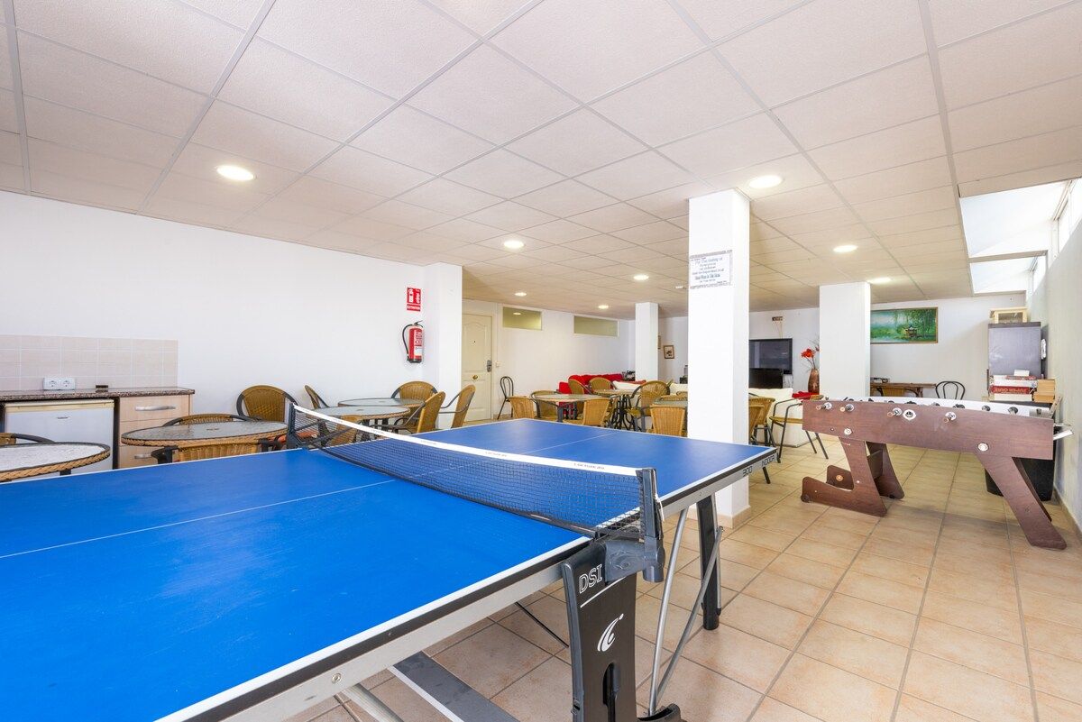 A room with a ping pong table, chairs, a foosball table, and a fire extinguisher on the wall.