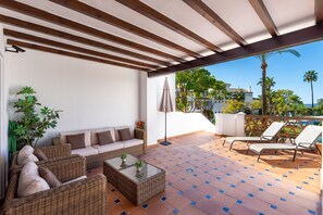 Une terrasse extérieure aménagée avec des meubles en osier, une table basse en verre et une vue sur une piscine et des palmiers.