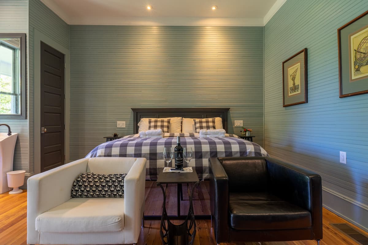 A bedroom with a checkered bedspread, a small table with a bottle and glasses, a white armchair, and a black leather chair.