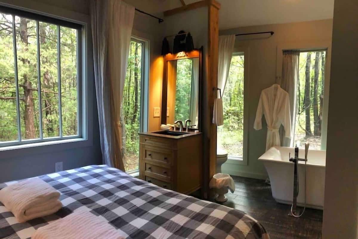 A bedroom with a checkered bedspread, a wooden dresser, a mirror, and a view of the outdoors through large windows.