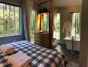 Un dormitorio con una colcha a cuadros, un aparador de madera, un espejo y vistas al exterior a través de amplios ventanales.